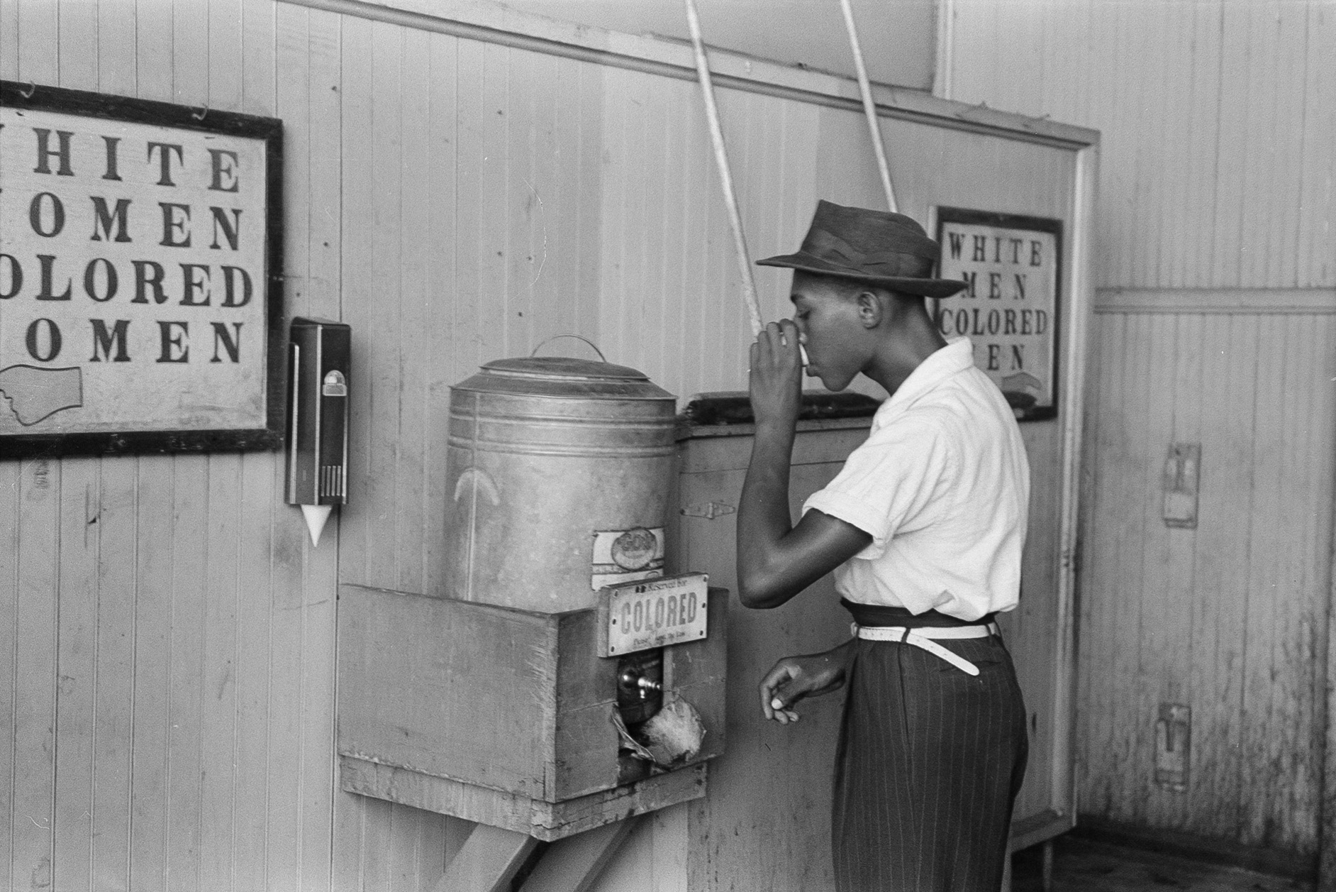 A Black man drinking from a water cooler marked “Colored” in a streetcar terminal in Oklahoma City, Oklahoma.